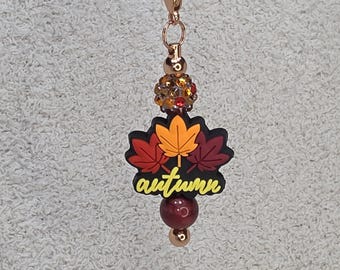 Op de afbeelding: Een roségouden bedel met een karabijnsluiting, met een cluster van oranje en rode strass-steentjes, een zwart en geel "herfst" bladontwerp en een bordeauxrode kraal. De bedel is ontworpen om aan een ketting of armband te worden bevestigd.