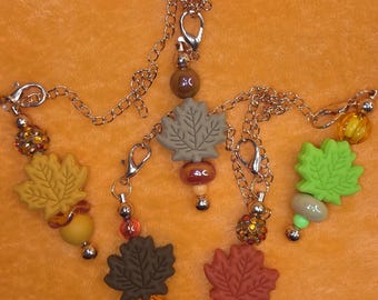Op de afbeelding: Een verzameling herfstthema-bedels met blad- en kralenontwerpen. Elke bedel heeft een blad in tinten geel, groen en bruin, gecombineerd met decoratieve kralen en een ketting. De achtergrond is effen oranje.