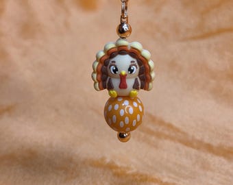 Op de afbeelding: Een bedel met een cartoon kalkoen ontwerp. De kalkoen heeft een wit gezicht, bruine veren en een rode lellen. Hij zit op een gele kraal met witte stippen, hangend aan een goudkleurige ketting.