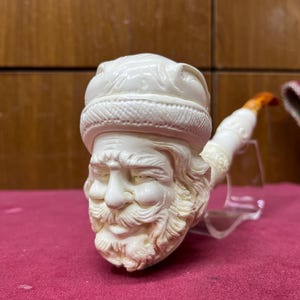 May include: A carved ivory-colored tobacco pipe shaped like a bearded man's head, wearing a hat. The pipe's stem is white with an orange tip. The pipe is displayed on a clear stand against a red background.