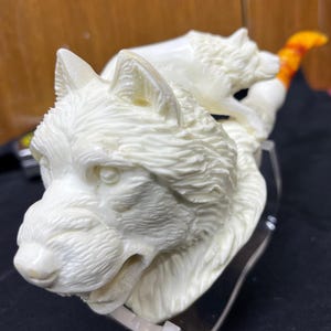 May include: Ivory-colored sculpture of two wolves. The larger wolf is in the foreground, with detailed fur texture and a slightly open mouth. A smaller wolf is positioned behind it. The tail has orange and yellow accents. The sculpture is mounted on a clear base.