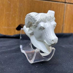 May include: A white ram sculpture with detailed horns and facial features. The ram is mounted on a clear acrylic stand, showcasing the sculpture's design. The sculpture is set against a dark background.