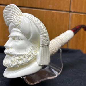 Può includere: Pipa da tabacco color avorio con un'intagliatura dettagliata del volto di un uomo con un turbante. La pipa ha un gambo curvo con bocchino rosso scuro e bianco. La testa è scolpita con dettagli intricati, che mostrano barba e lineamenti.