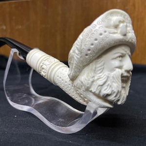 May include: Ivory-colored tobacco pipe with a detailed carving of a bearded man wearing a hat. The pipe has a black stem and is displayed on a clear stand. The bowl and stem are intricately decorated with patterns.