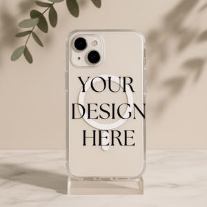 Op de afbeelding: Transparante telefoonhoes met de tekst "YOUR DESIGN HERE". De hoes heeft een wit cirkelvormig ontwerp en wordt gepresenteerd op een transparante standaard. De telefoonhoes heeft twee cameralenzen.