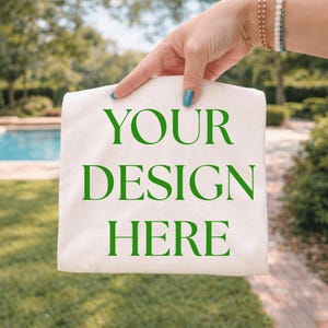 May include: A folded, off-white fabric item with the words "YOUR DESIGN HERE" printed in green. The item is held up against a blurred outdoor background of green grass, trees, and a swimming pool. The person holding the item has blue nail polish.