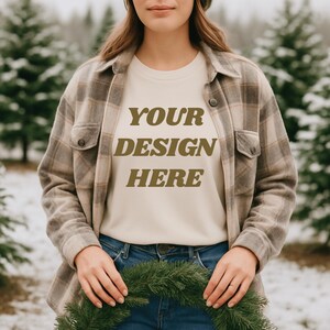 May include: A person wearing a beige t-shirt with the text "YOUR DESIGN HERE" in brown letters, paired with a plaid button-up shirt and blue jeans. The person is holding a green wreath. The background features snow-covered evergreen trees.