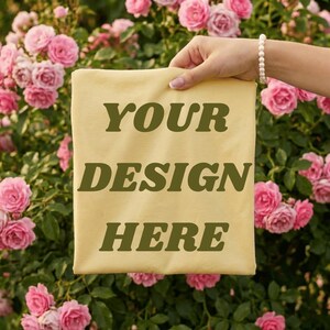 May include: A light yellow t-shirt featuring the dark olive green text "YOUR DESIGN HERE" printed in a bold, serif font. The shirt is held up against a background of blooming pink roses, showcasing the fabric color and the clear, centered lettering.