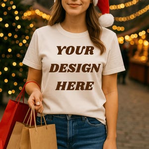 May include: Cream-colored t-shirt with the text "YOUR DESIGN HERE" in brown letters. The person is wearing a Santa hat and holding shopping bags. The background features Christmas lights.