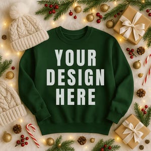 May include: A dark green sweatshirt with the text "YOUR DESIGN HERE" in white. The sweatshirt is surrounded by holiday decorations, including a cream-colored knit hat, wrapped gifts, ornaments, pine cones, and candy canes.