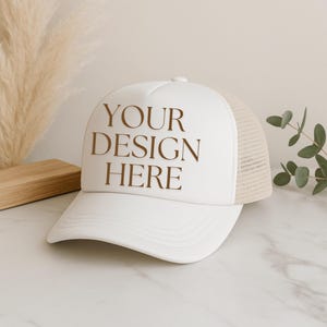 Puede incluir: Gorra de camionero blanca con visera curva y parte trasera de malla. El panel frontal muestra el texto "YOUR DESIGN HERE" en marrón. Accesorio neutro.