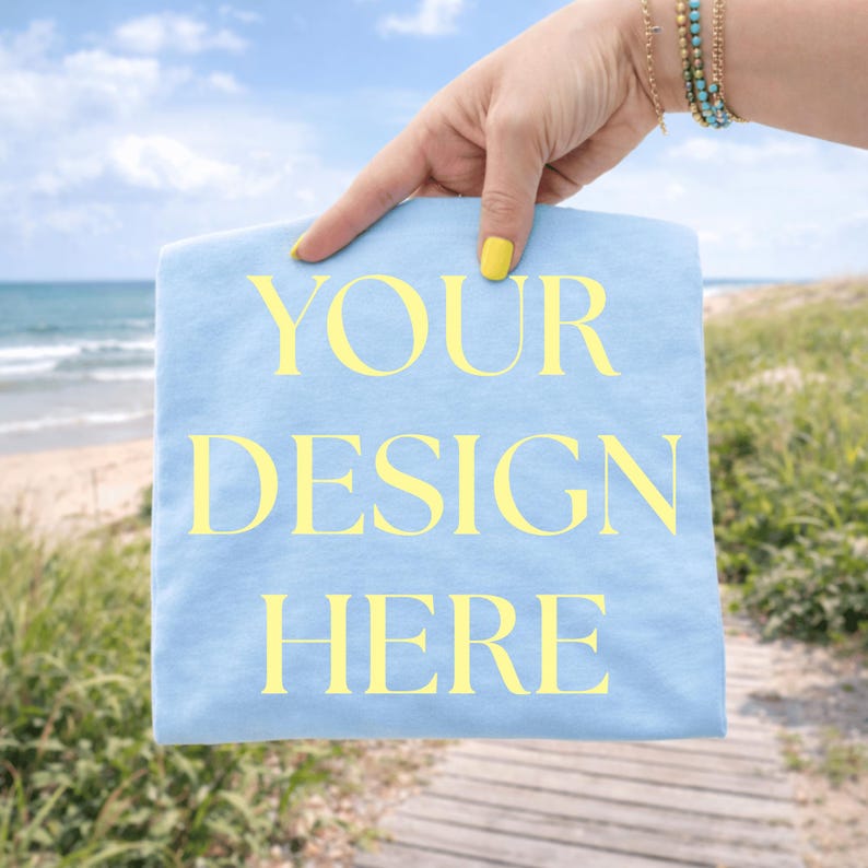 Light blue folded T-shirt mockup held by hand with yellow nails and bracelets outdoors near the beach, blank shirt mockup with no collar visible
