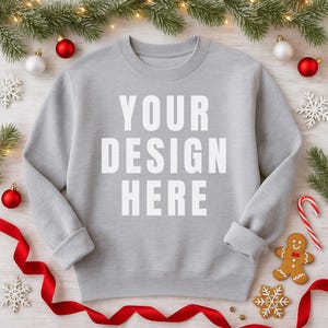 May include: A light gray sweatshirt with the text "YOUR DESIGN HERE" in white block letters. The sweatshirt is surrounded by Christmas decorations, including red and white ornaments, snowflakes, a gingerbread man, and a candy cane.