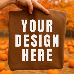 May include: A brown tote bag with the text "YOUR DESIGN HERE" in white. The bag is held up in front of a pumpkin patch with orange pumpkins and fall foliage in the background.