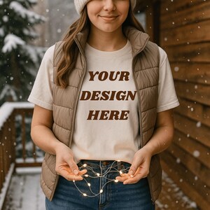 May include: A cream-colored t-shirt with the text "YOUR DESIGN HERE" in brown, worn with a tan vest and blue jeans. The person is holding a string of small, warm white lights. The background includes a snowy outdoor scene and a wooden wall.