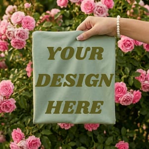 May include: A light green t-shirt with the text "YOUR DESIGN HERE" in a darker green font. The t-shirt is held up in front of a background of pink roses.