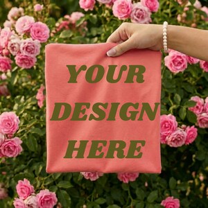 May include: A coral-colored t-shirt with the text "YOUR DESIGN HERE" in a dark green font. The shirt is held up in front of a background of pink roses.