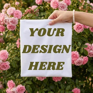 May include: A white t-shirt with the text "YOUR DESIGN HERE" in a bold, olive green font. The shirt is held up against a backdrop of pink roses and green foliage.