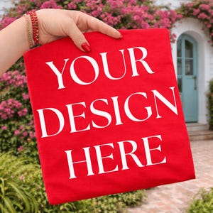 May include: A red t-shirt with the words "YOUR DESIGN HERE" printed in white. The shirt is held up in front of a background of pink flowers and a blue door.