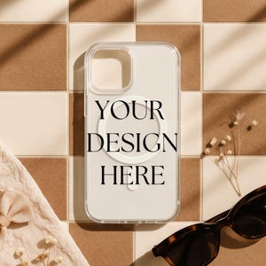 May include: Clear phone case with the text "YOUR DESIGN HERE" in black. The case has a white circular design on the back. The case is on a brown and beige tiled surface, with sunglasses and a bow in the background.