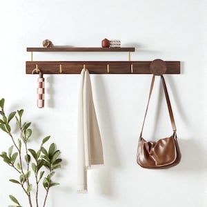 Puede incluir: Un estante y sistema de ganchos de pared de madera. La madera marrón oscuro tiene detalles y ganchos de color latón. Un bolso marrón claro cuelga de un gancho, y una toalla color crema cuelga de otro. Un paraguas decorativo y objetos pequeños están en el estante.