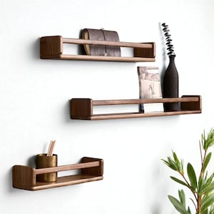 May include: Three wooden floating shelves on a white wall. The shelves are made of dark wood with a simple design. One shelf holds a brown leather notebook, another a book and a dark vase, and the third a small brass cup. Ideal for home decor.