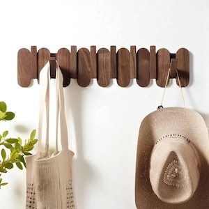 Könnte beinhalten: Ein hölzerner, an der Wand befestigter Kleiderständer mit einer Reihe von vertikalen und horizontalen Holzstücken. Eine beige Tragetasche und ein Strohhut hängen an dem Ständer. Der Ständer ist an einer weißen Wand befestigt.