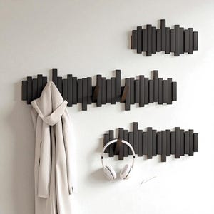 Könnte beinhalten: Drei dunkelgraue Holz-Kleiderhaken an einer weißen Wand. Jeder Haken hat eine Reihe vertikaler Holzstücke. Ein Haken hält einen weißen Mantel, ein anderer weiße Kopfhörer, und der dritte ist leer.