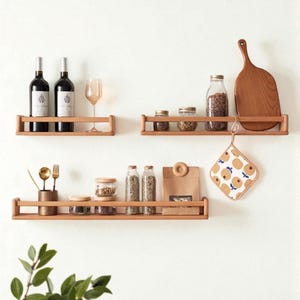 May include: Three wooden shelves mounted on a white wall, each holding various kitchen items. The top shelf displays wine bottles and a glass. The middle shelf has jars of spices and a cutting board. The bottom shelf holds utensils and more jars.