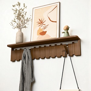 Peut inclure: Une étagère murale en bois avec un porte-manteau intégré. L'étagère contient une impression d'art abstrait encadrée, un vase avec des fleurs séchées et une petite figurine. Une écharpe à motifs noir et blanc et un sac à main blanc sont suspendus au porte-manteau.