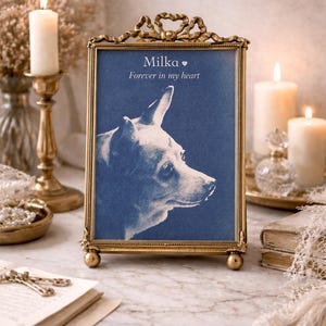 May include: A framed portrait of a dog named Milka, with the text "Forever in my heart." The portrait is in a gold-colored ornate frame with a blue background. The image is set on a marble surface with candles and decorative items.