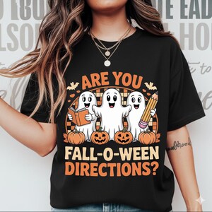 Puede incluir: Camiseta negra con un diseño temático de Halloween. El diseño presenta tres fantasmas de dibujos animados, calabazas y el texto "ARE YOU FALL-O-WEEN DIRECTIONS?" en naranja y blanco. Los fantasmas sostienen un libro y un lápiz.