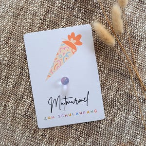 Könnte beinhalten: Eine weiße Karte mit einer bunten Illustration einer Schultüte und dem Schriftzug "Mutmurmel ZUM SCHULANFANG" in schwarzer Schrift. Eine kleine, irisierende Reißzwecke ist an der Karte befestigt. Die Karte liegt auf einer strukturierten, neutralfarbenen Oberfläche mit Trockenblumen.