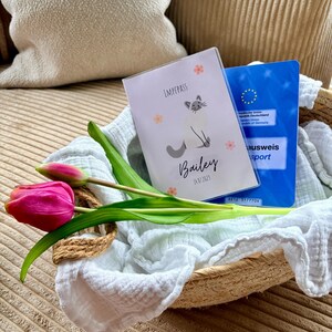 Könnte beinhalten: Ein geflochtener Korb enthält eine rosa Tulpe, ein weißes Tuch und zwei Dokumente. Ein Dokument hat eine Katzenillustration und den Namen "Bailey". Das andere ist ein blaues Dokument mit Text. Der Korb liegt auf einer strukturierten Oberfläche.