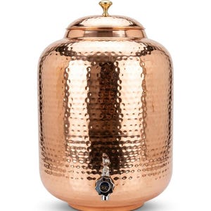 Puede incluir: Un gran dispensador de agua cilíndrico de cobre que presenta una textura martillada distintiva en toda su superficie. El recipiente incluye una tapa extraíble con un pequeño pomo redondeado de color latón en la parte superior y un grifo de metal plateado.