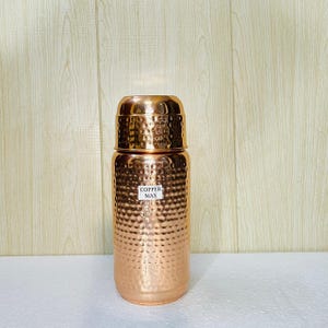 Könnte beinhalten: Eine kupferfarbene Wasserflasche mit strukturierter, gehammerter Oberfläche. Die Flasche hat einen passenden Deckel und ein Etikett mit der Aufschrift "COPPER MAX". Die Flasche ist zylindrisch und steht auf einer weißen Oberfläche.