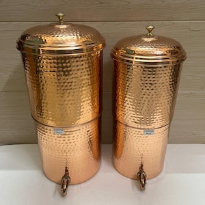 May include: Two hammered copper water dispensers with lids and spigots. Each cylindrical vessel features a polished metallic finish and a small rectangular label on the front that reads COPPER MAX in black text. The containers stand upright.