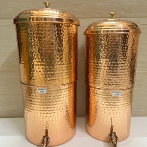 May include: Two copper water dispensers with a hammered texture. Each dispenser has a lid, a spigot, and a small label that reads "COPPER MAX". The dispensers are cylindrical and have a shiny, metallic finish.