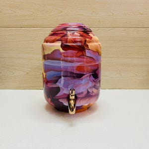 May include: A cylindrical beverage dispenser featuring a vibrant, abstract floral pattern in shades of deep red, orange, purple, and soft lavender. The container has a rounded top lid and a metallic gold-toned spigot centred on the front.