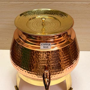 May include: A copper beverage dispenser with a hammered texture and a brass lid and base. The dispenser has a spigot and the text "COPPER MAX" on the front. The lid is round and has a small handle.