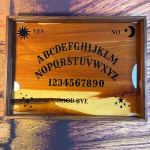 May include: A wooden Ouija board with a dark brown frame. The board features the alphabet, numbers, and the words "YES", "NO", and "GOOD BYE" in black. A sun and moon graphic are also present.