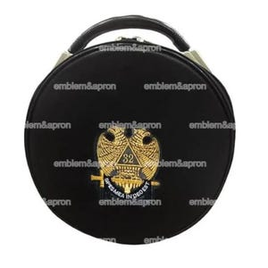Puede incluir: Estuche redondo negro con asa y emblema dorado. El emblema presenta un águila bicéfala, el número 32 y el texto "SPES MEA IN DEO EST". La funda tiene el texto "emblem&apron" repetido alrededor del borde.