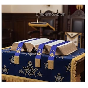 May include: Two open books with blue ribbons and gold fringe sit on a blue cloth with gold embroidery. The cloth is on a table with gold fringe. The text "Emblem&Apron" is visible.