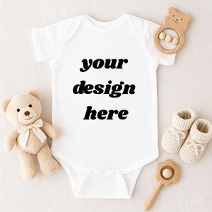 May include: A white baby onesie with the text "your design here" in black. The onesie is surrounded by a beige teddy bear, wooden toys, and knitted baby booties. A charming baby gift set.