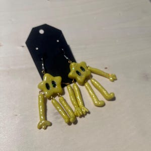 May include: A pair of yellow star-shaped earrings with attached arms and legs, featuring black eyes and a glittery finish. The earrings are displayed on a black card, showcasing a dangling design with articulated limbs extending from the star bodies.