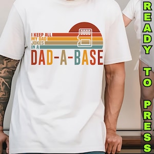May include: White t-shirt with the text "I KEEP ALL MY DAD JOKES IN A DAD-A-BASE" in a retro design. The design includes a sunset graphic with a memory card icon. The words "READY TO PRESS" are printed vertically on the right side.