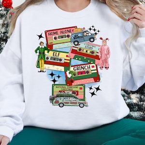 May include: White sweatshirt featuring a colorful graphic of Christmas-themed cassette tapes. The tapes display titles like "Home Alone," "Elf," and "Grinch." Additional elements include a car with a Christmas tree and cartoon characters.