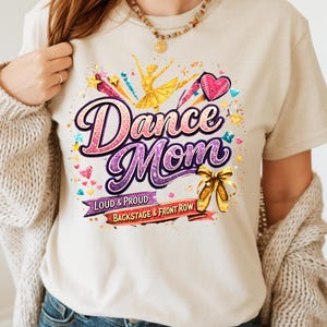 May include: Beige t-shirt with a colorful "Dance Mom" graphic. The design includes a ballerina, hearts, stars, and ballet shoes. Text reads "Loud & Proud" and "Backstage & Front Row".