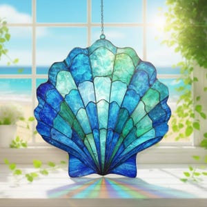 Pode incluir: Um apanhador de sol de concha de vidro manchado em tons de azul e verde pendurado em uma corrente. A concha está em frente a uma janela com vista para o oceano e o céu. O apanhador de sol é um item decorativo.