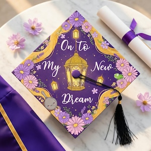 May include: A purple graduation cap with the words "On to My New Dream" in white script, surrounded by floral and lantern designs. A black tassel hangs from the cap. A rolled diploma with a purple ribbon is also present.